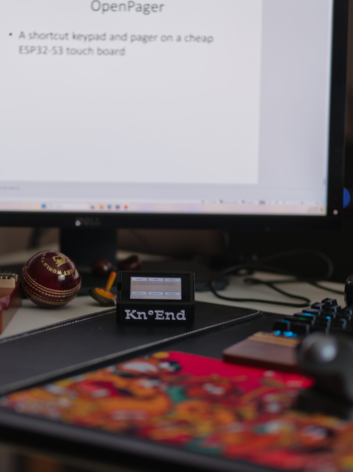 The Kn°End board in its black printed case on a desk in front of a monitor showing a slide. The board's screen shows the slide's speaker notes with Prev, End, Next, First, Black and White keys.